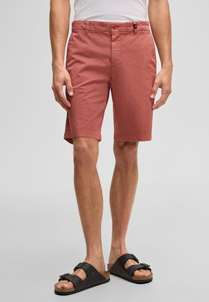 Man draagt zalmkleurige knielange shorts, wit shirt en zwarte sandalen met dubbele bandjes, staat tegen een effen achtergrond.