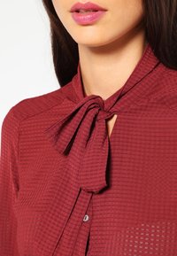Blouse bordeaux en tissu texturé avec un lien au niveau du col. Présente une fermeture à boutons et des motifs subtils à rayures diagonales.