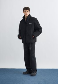 Junger Mann steht und trägt eine schwarze Steppjacke, schwarze zerrissene Jeans mit Mustern und schwarze Turnschuhe auf blauem Teppich.