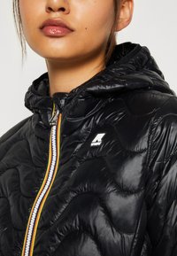 Giacca a vento nera con motivo trapuntato, cappuccio e zip multicolore. Presenta un logo sul petto e una texture lucida.