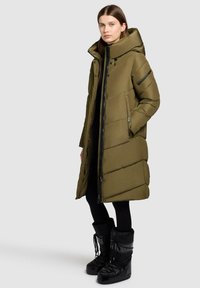 Kvinna som bär en lång olivgrön vadderad kappa med stor huva, svarta leggings och svarta vinterstövlar som står mot en enfärgad bakgrund.