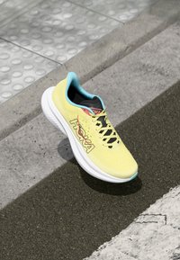 Baskets de course jaune vif avec tige en mesh, accents bleus, lacets noirs et semelle intermédiaire blanche. Présence du logo "Hoka" affiché en évidence.