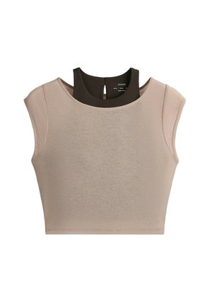 Femme debout portant un haut court sans manches beige, des leggings foncés taille haute, des chaussettes blanches et des baskets beiges sur un fond gris.