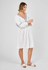 Weißes, knielanges Kleid mit V-Ausschnitt und blauer Blumenstickerei an den Ärmeln. Leichter Stoff mit strukturiertem Finish, kombiniert mit flachen Sandalen.