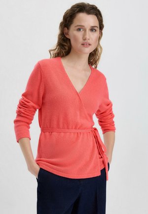 Korallfarbener Wickelpullover aus weichem Strickmaterial mit V-Ausschnitt, langen Ärmeln und gebundenem Taillenband; kombiniert mit dunklen Hosen.
