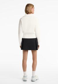White, knitted sweater with a wide collar, paired with a black pleated skirt. Model wears white socks and light sneakers. Plain background.