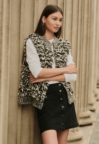 Gestructureerd zwart-wit mouwloos vest over een wit langemouwen shirt, gecombineerd met een zwarte denim rok met voorknopen.