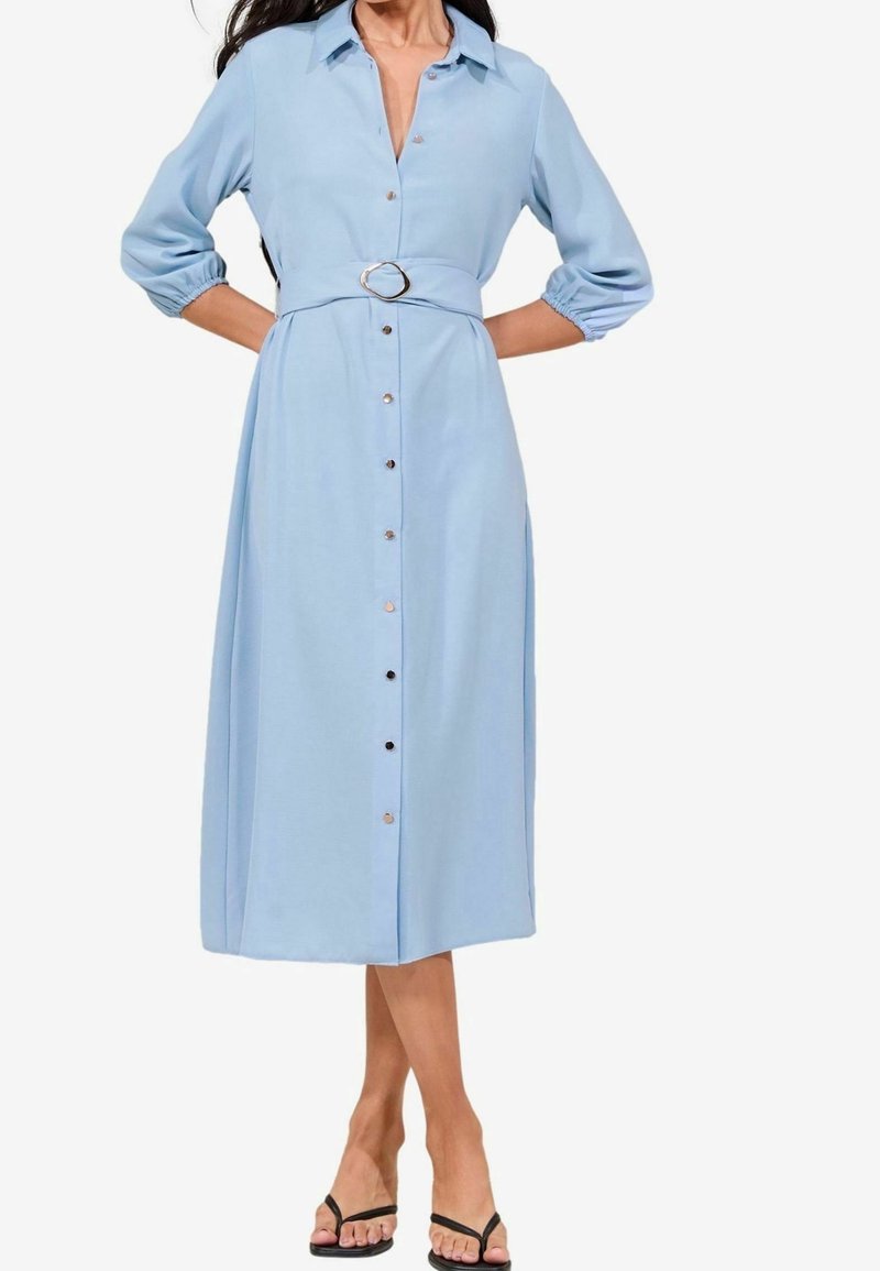 Femme portant une robe chemise midi bleu clair avec des boutons, une taille ceinturée, des manches trois-quarts et des sandales noires à bout ouvert.