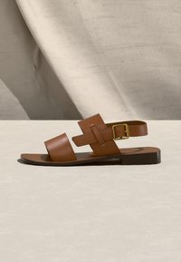 Καφέ δερμάτινο φλατ πέδιλο με δύο φαρδιές λουριά και χρυσή αγκράφα στον αστράγαλο, σε ανοικτή υφασμάτινη επιφάνεια.