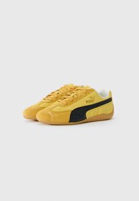 Zapatillas de suede amarillas de Puma con detalles de franjas negras, cordones y suelas de goma, colocadas lado a lado sobre un fondo blanco.