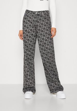 Persona vistiendo jeans grises holgados con un patrón geométrico "KL" por todo el tejido y zapatillas blancas, de pie contra un fondo blanco liso.