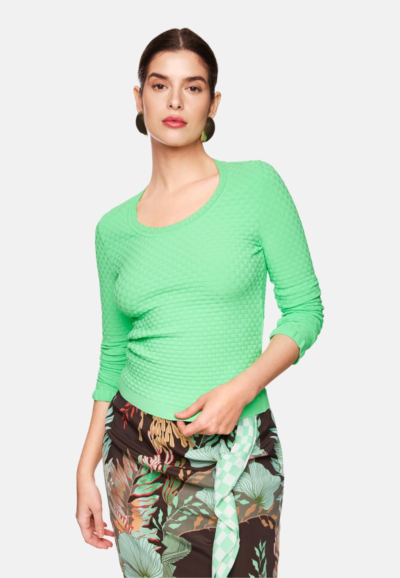 Grüner, strukturierter Langarmoberteil mit rundem Ausschnitt, kombiniert mit einem gemusterten Rock mit floralen Designs und Farbkontrasten.