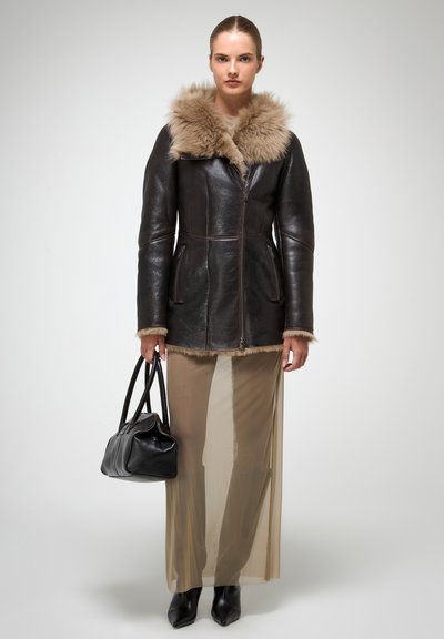 Brūna ādas jaka ar beigu krāsas kažokādu, ar rāvējslēdzēju priekšā un sānu kabatām. Valkāta ar caurspīdīgu garu svārku un melnu rokassomu.