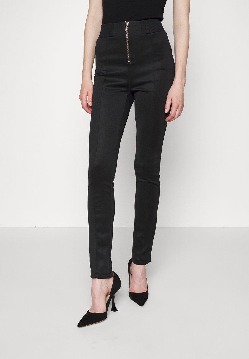 Black stretchy trousers with a fitted design, featuring a front zip closure and smooth texture. Paired with black pointed-toe heels.
