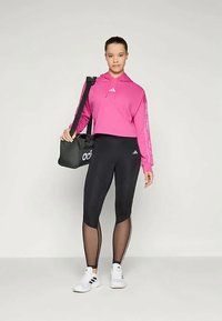 Ροζ κοντό φούτερ με λευκές λεπτομέρειες λογοτύπου, μαύρες κολάν με λεπτομέρειες από δίχτυ, και λευκά αθλητικά παπούτσια. Κρατάει μία μαύρη τσάντα.