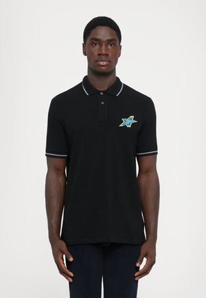 Man wearing black polo shirt with striped collar and sleeve edges, featuring a blue and green star emblem on the chest, standing against white background.