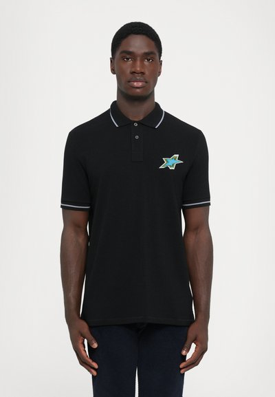 Mand iført sort polo-shirt med stribet krave og ærmekanter, prydet med et blåt og grønt stjernesymbol på brystet, stående foran en hvid baggrund.