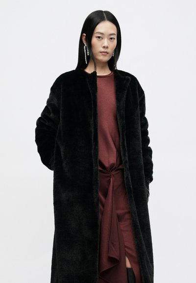 Crni kaput od umjetnog krzna s opuštenim krojem i otvorenim prednjim dijelom. Model nosi bordo haljinu s čvorastim detaljem i dugim crnim rukavima.