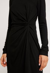 Svart långärmad klänning med en vriden knutdetalj vid midjan, gjord av mjuk jersey med draperad textur.