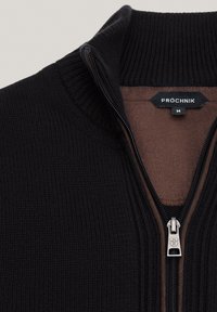 Czarny sweter z ribbowanym splotem, zapinany na zamek błyskawiczny, z wysokim kołnierzem, posiadający srebrny zamek oraz kontrastową brązową podszewkę. Na widoku etykieta marki.