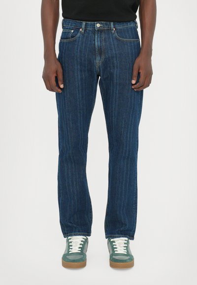 Jeans de mezclilla azul oscuro con líneas verticales tenues, diseño de cinco bolsillos y cierre de botón. Llevados con zapatillas verdes y cordones blancos.