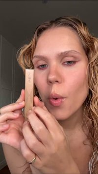 Gul rektangulär kosmetikprodukt som hålls i händerna, med upphöjda diagonala linjer. Modellen har blanka läppar och vågigt hår.