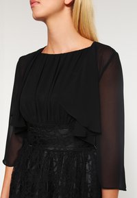 Czarna sukienka z plisowanym górnym panelem i przezroczystymi rękawami. Spódnica ozdobiona jest misternymi koronkowymi detalami. Gładka tekstura z dopasowaną linią talii.