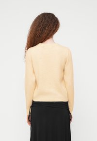 Femme aux longs cheveux bouclés portant un pull beige et une jupe noire, vue de dos sur un fond clair uni.