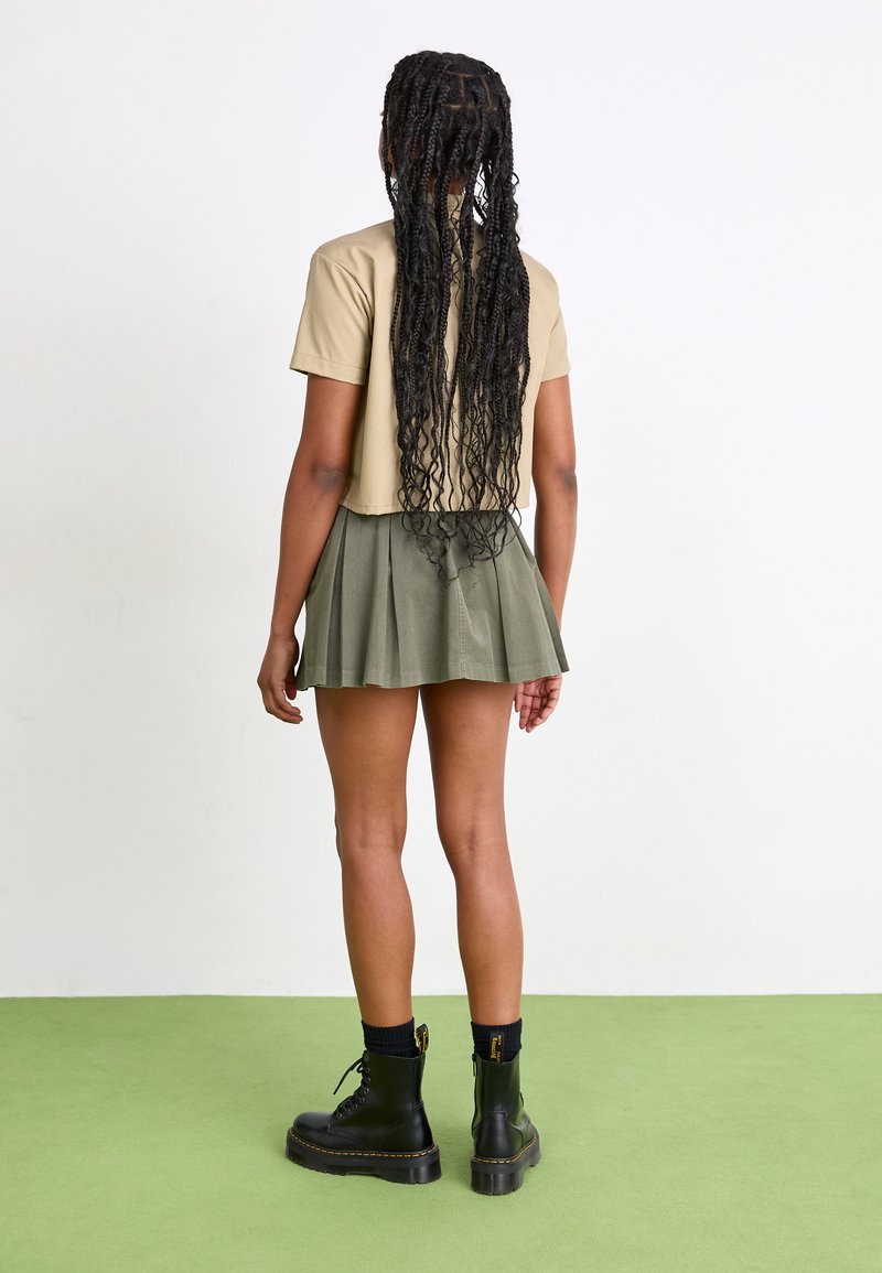 Tan gekort t-shirt met korte mouwen, gecombineerd met een groene geplooide mini-rok. Zwarte veterschoenen tot de enkel met gele stiksels. Groene vloer.