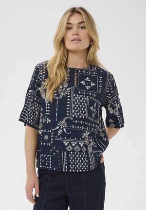 Mujer joven con el pelo largo y rubio, vestida con una blusa azul marino con estampado y pantalones oscuros, de pie frente a un fondo liso.
