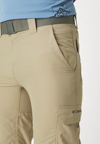 Pantalons cargo beiges en tissu léger avec des poches latérales, des coutures visibles et une coupe décontractée, complétés par une ceinture tissée grise.