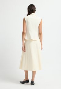 Cremefarvet ærmeløs blazer med en tætsiddende pasform, kombineret med en knælængde nederdel i matchende stof og sorte spidse sko.