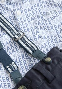 Biała koszula z powtarzającym się niebieskim nadrukiem "BAKER", zestawiona z ciemnoniebieskimi szelkami o teksturowanym splocie i srebrnym osprzętem.
