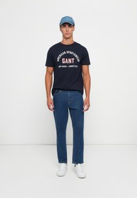 Tricou bleu marin cu imprimeu grafic alb și roșu; asortat cu blugi denim de culoare bleu închis și adidași albi; șapcă bleu deschis.