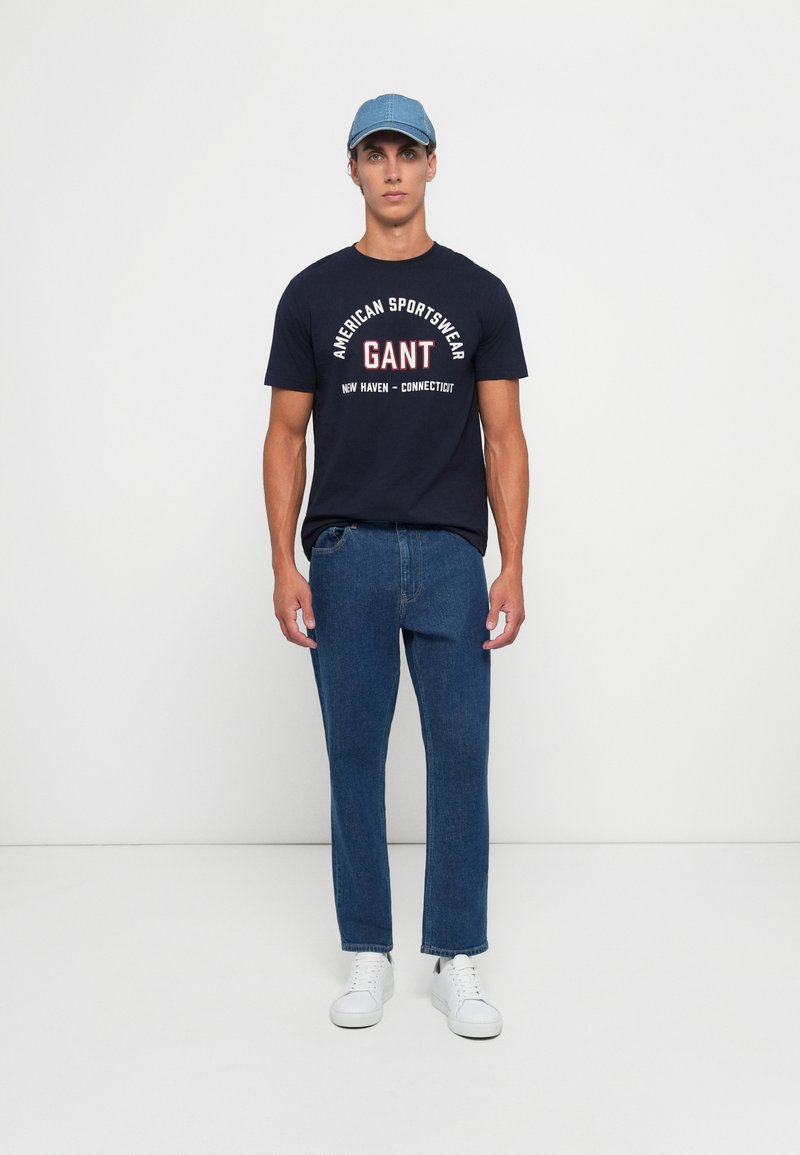 Granatowa koszulka z białym i czerwonym nadrukiem graficznym; zestawiona z ciemnoniebieskimi dżinsami i białymi sneakersami; jasnoniebieska czapka.