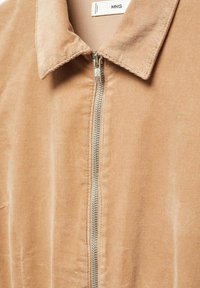 Veste en velours côtelé beige avec une fermeture éclair sur le devant et un col. Présente une texture à côtes verticales avec des éléments en métal argenté sur la fermeture éclair.