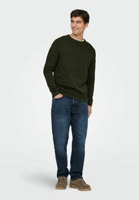 Suéter texturizado verde oscuro con cuello redondo, combinado con vaqueros de mezclilla azules y zapatos beige. El modelo está de pie con las manos en los bolsillos.