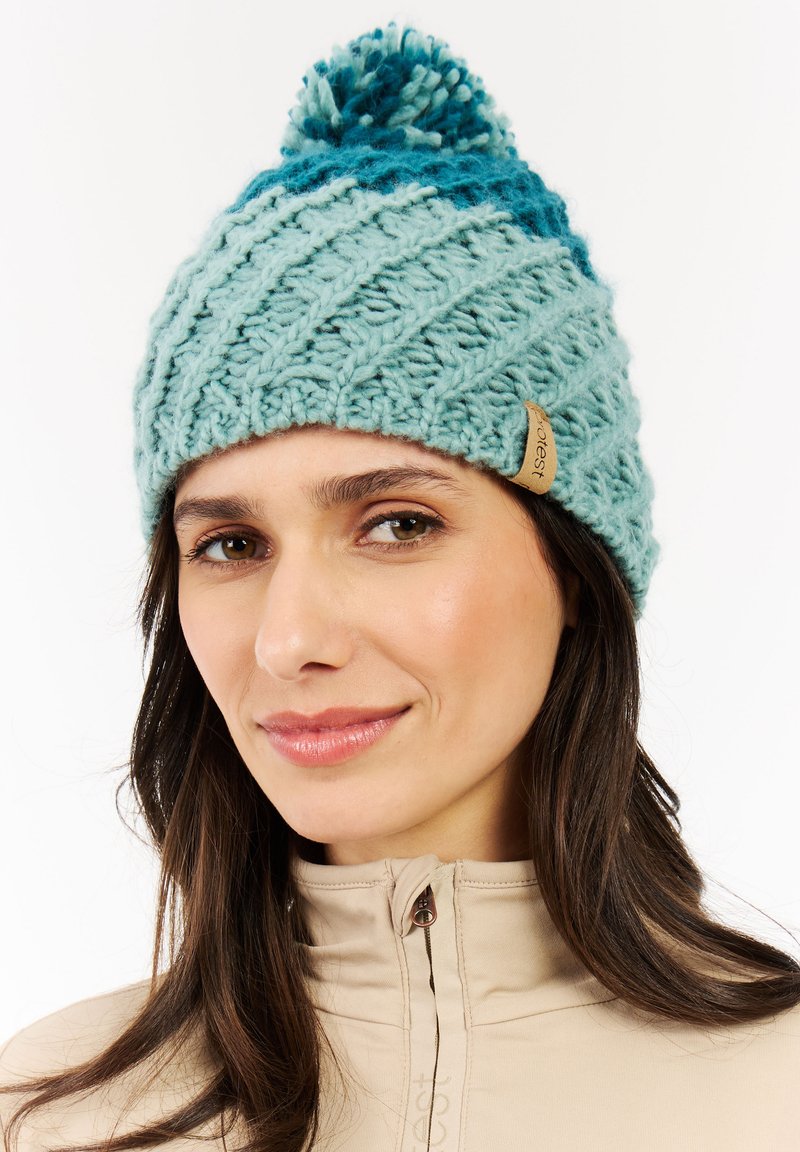 Lichtblauwe gebreide beanie met een gestructureerd diagonale patroon, voorzien van een pompon bovenop en een bruin leren logo-tag aan de zijkant.