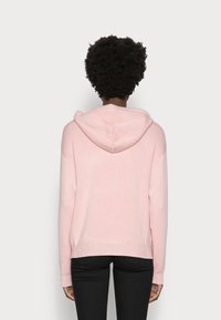 Persona con capelli ricci corti che indossa un maglione con cappuccio rosa chiaro e pantaloni neri, in piedi rivolta di spalle su uno sfondo neutro.