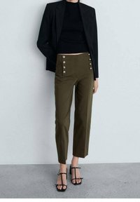 Vrouw in een zwarte cropped blazer en top, olijfgroene hoge taillebroek met dubbele rijen decoratieve knopen, en zwarte haksandalen.