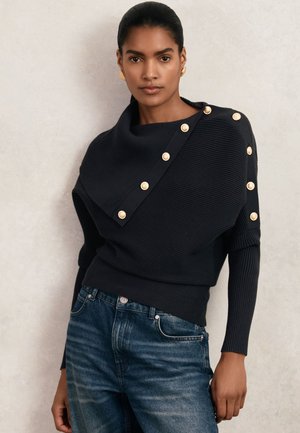 Donna che indossa un maglione nero a coste con bottoni dorati lungo un colletto e maniche asimmetriche, abbinato a jeans blu.