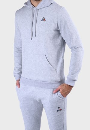 Mężczyzna w jasno szarej bluzie z kapturem i pasujących spodniach dresowych z logo Le Coq Sportif, ręce w kieszeniach, stoi na jednolitym tle.