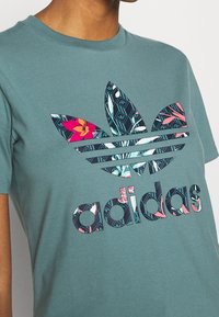 Teal bomullströja med en stor blommig och geometrisk mönstrad Adidas-logotyp i flera färger, med korta ärmar och rund halsringning.