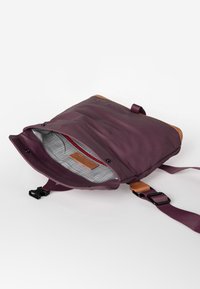 Lila Tasche mit glatter Oberfläche, braunen Lederakzenten, verstellbarem Riemen und einem grauem, gemusterten Innenraum, der eine rote Reißverschlusstasche enthält.
