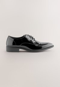 Chaussure de ville pour homme en cuir verni noir avec laçage fermé, petit talon et finition polie, vue de côté sur fond neutre.