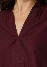 Zbli�żenie na osobę noszącą głęboki burgundowy T-shirt z dekoltem w serek, z delikatnymi pionowymi przeszyciami wokół dekoltu.