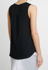 Black sleeveless top with a rounded back, pleated detail, and curved hem. Made from a smooth fabric, paired with a silver skirt.