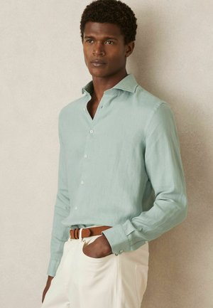 Hellgrünes Leinenhemd mit Button-Down-Kragen, langen Ärmeln und Knöpfen. Kombiniert mit beigen Hosen und einem braunen Gürtel. Glatte Textur.