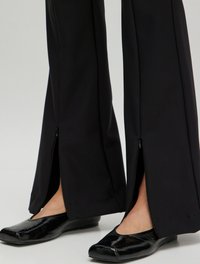 Schwarze weit geschnittene Hosen mit Reißverschlüssen an den Knöcheln werden mit glänzend schwarzen Ballerinas kombiniert, was ein elegantes, schlankes Design betont.
