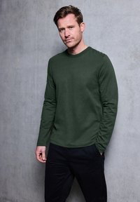 Zielona oliwkowa koszulka z długim rękawem, wykonana z miękkiego materiału, z okrągłym dekoltem i subtelnym logo na piersi, w połączeniu z ciemnymi spodniami.